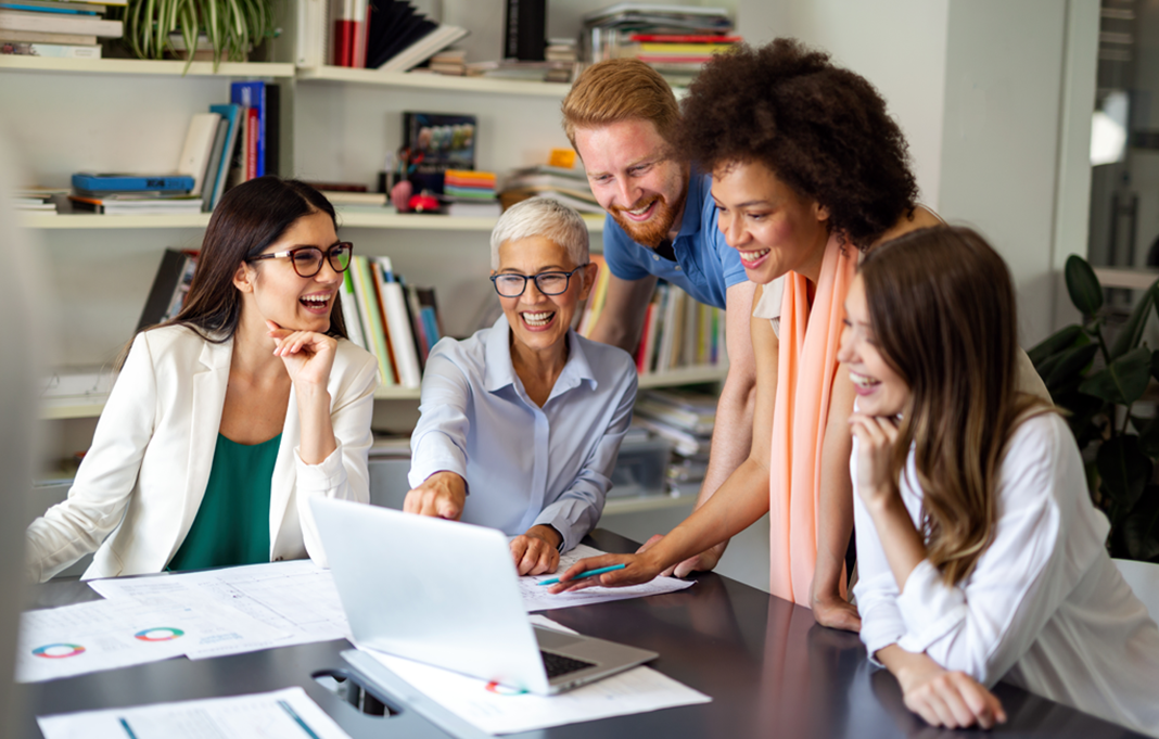 Group of five people around a laptop in an office smiling and discussing work